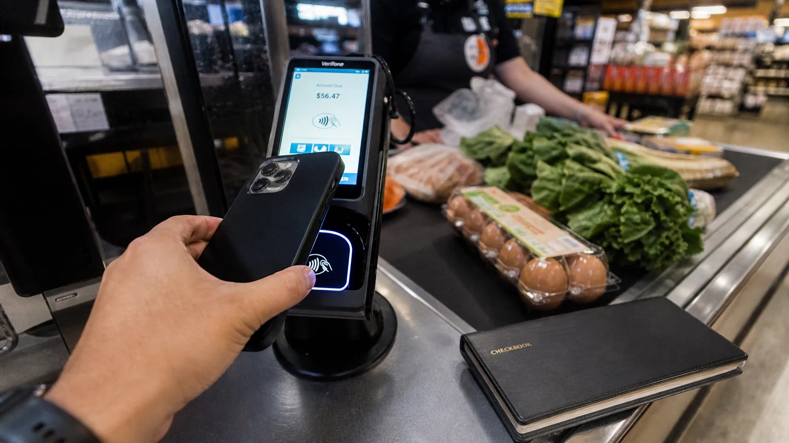 A close-up shot of a customer tapping their phone to pay at a grocery store register, with a closed checkbook sitting nearby.