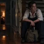 A weary woman in work clothes sitting on a wooden staircase holding keys with a duffel bag at her feet.