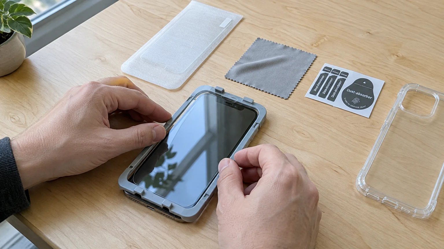 Hands applying a screen protector to a smartphone using an alignment frame on a wooden desk beside a clear case.