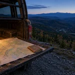 A map, compass, and book on an open vehicle tailgate overlooking a wide mountain valley at dusk.